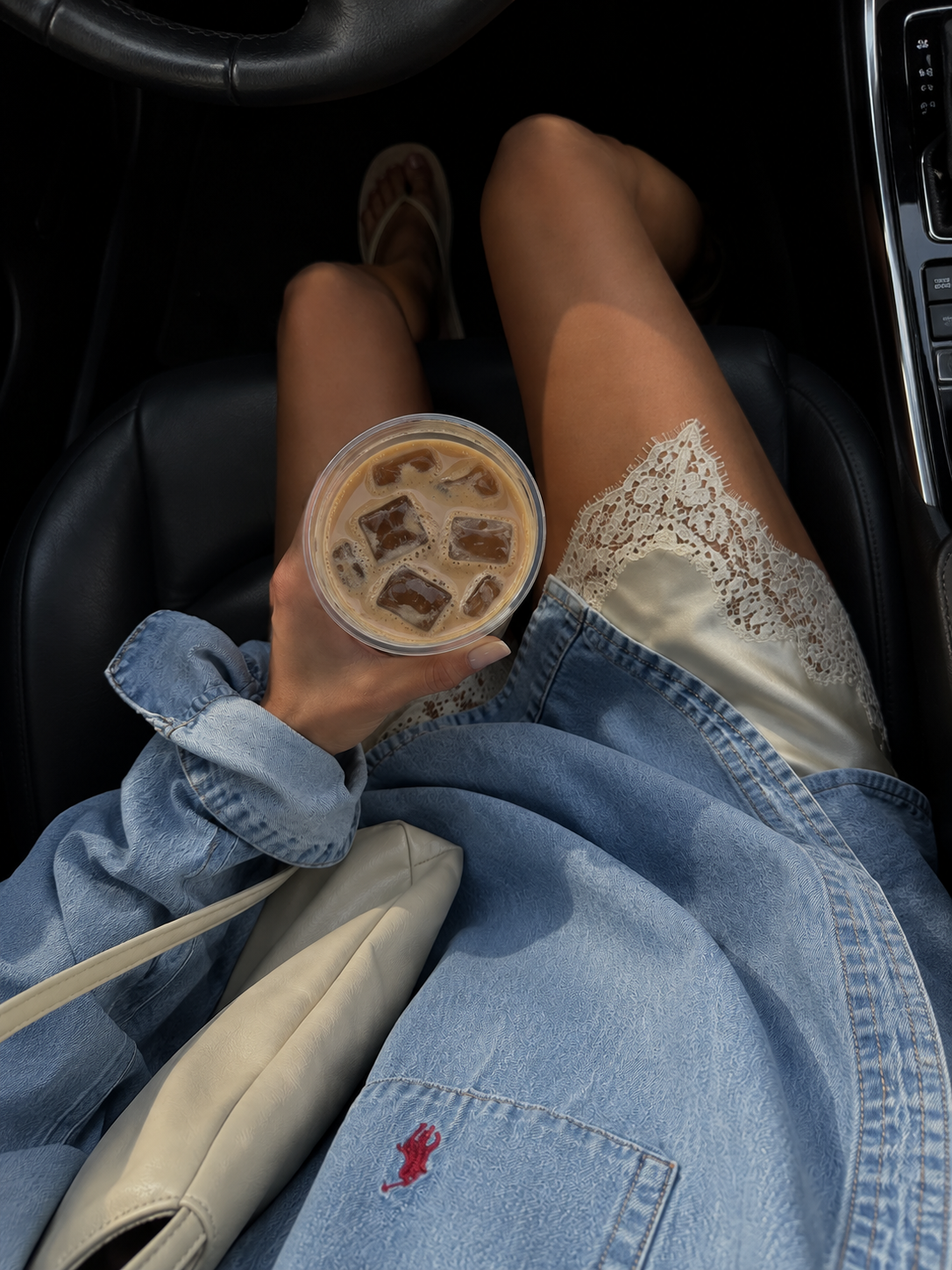 iced coffee held above an ivory lace slip and denim jacket in a car with black leather seats