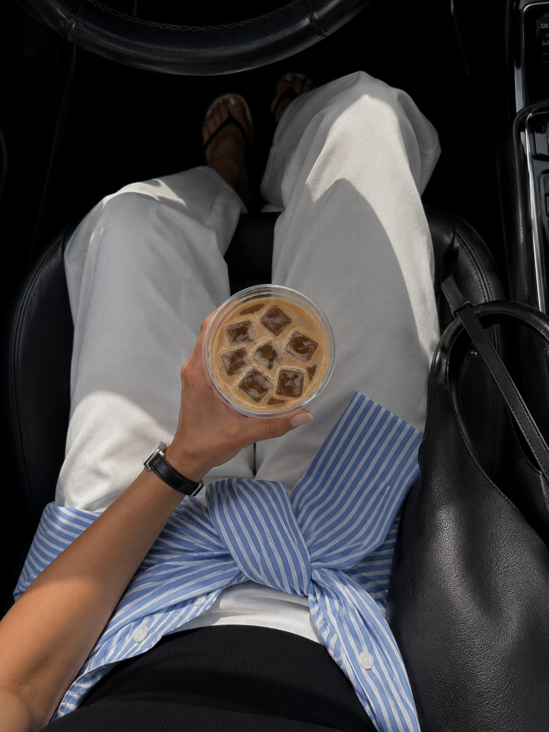 iced coffee held above ivory trousers and a blue striped shirt tied at the waist in a car with black leather seats