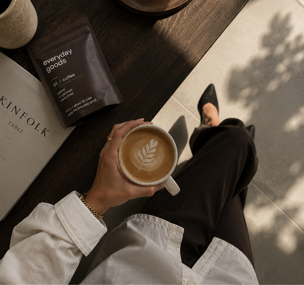 hand holding a latte with leaf art above dark trousers and black mules, with an everyday goods coffee pouch and Kinfolk book on a dark wood table