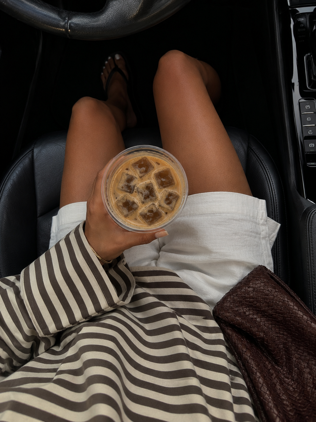 iced coffee held above white shorts and a striped shirt in a car with black leather seats